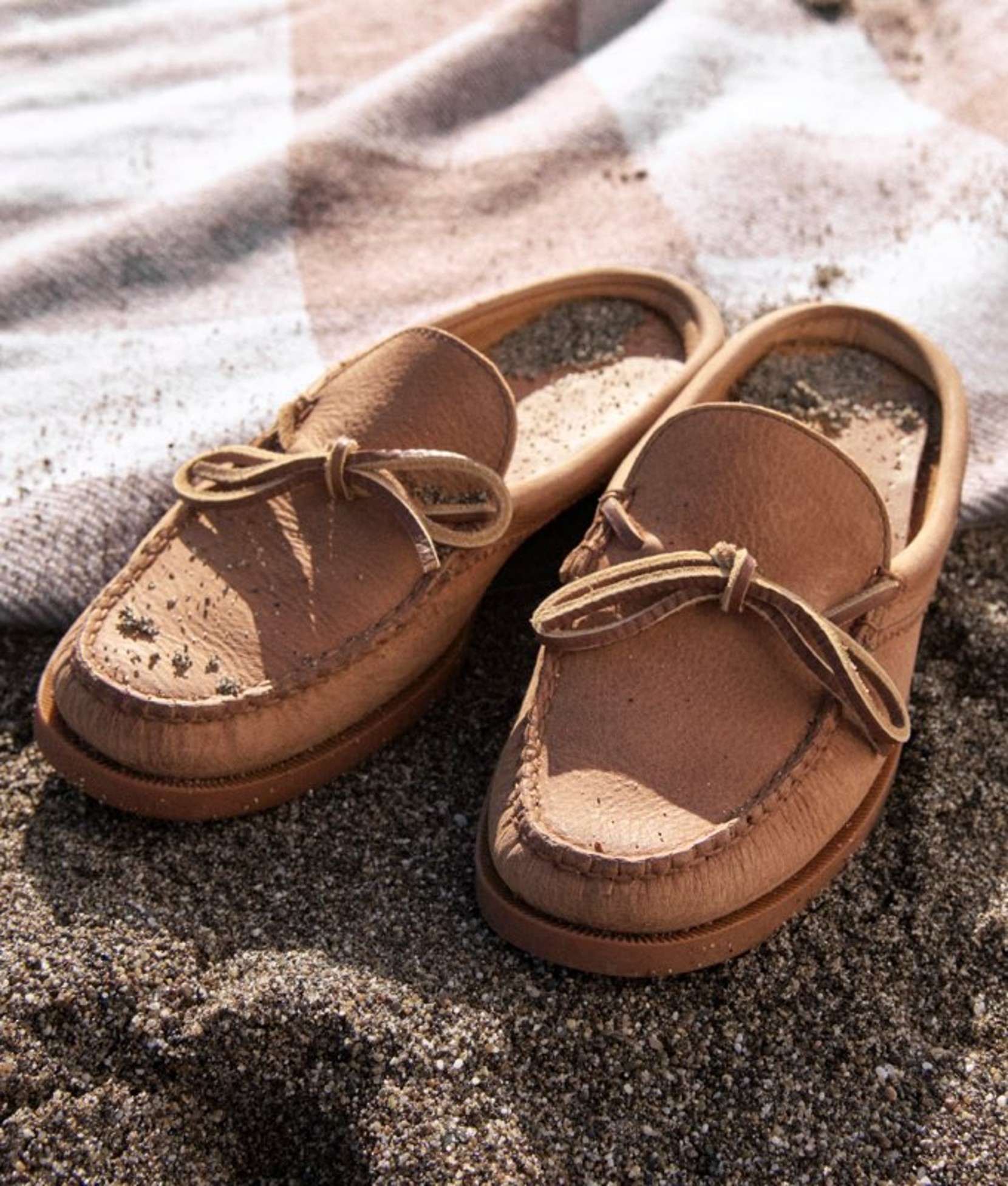 Pair of brown casual shoes placed on sand near the water’s edge. 