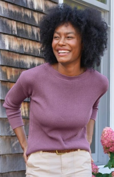 Person in mauve textured crewneck sweater paired with light beige pants stands in front of a rustic shingled wall.