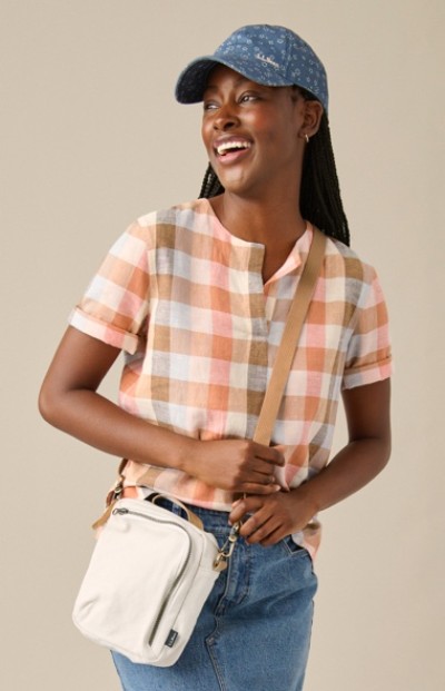 A woman in a blue cap, plaid shirt, and blue jeans, holding a white crossbody bag. The shirt has shades of orange, white, and beige.