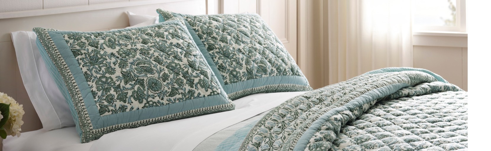 Neatly made bed with teal patterned pillows and quilt, soft lighting, and a bright, airy bedroom setting.