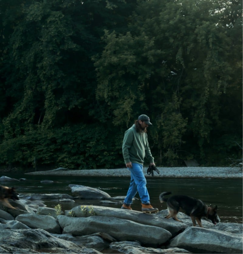 Noah Kahan wearing a green hoodie and L. L. Bean Boots walks on river rocks with his two dogs.