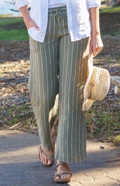 A woman wearing a white shirt and green striped pants, holding a straw hat, walking on a path.