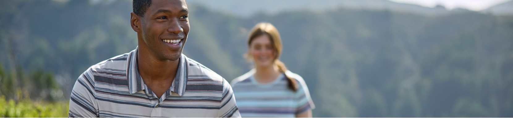 Two people hiking on a trail surrounded by greenery and hills in the background. Both are wearing striped shirts.