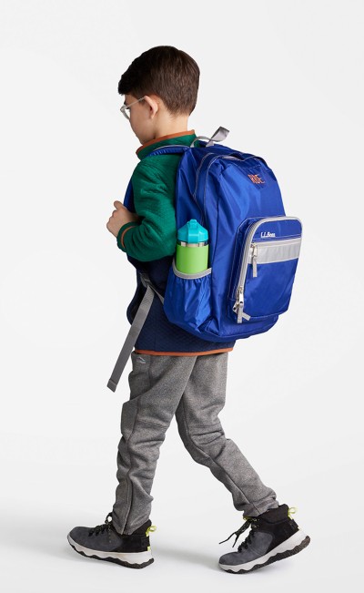 A young boy from slightly behind, wearing a medium blue school backpack.