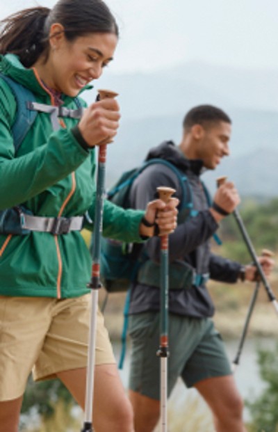 Two people using hiking poles while walking on a wooded trail with a lake in the background.