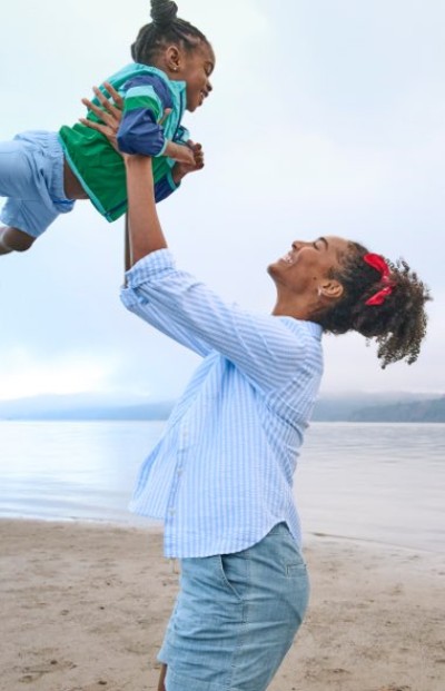 An adult lifts a child into the air near a calm lakeshore, with hills and water stretching across the background.