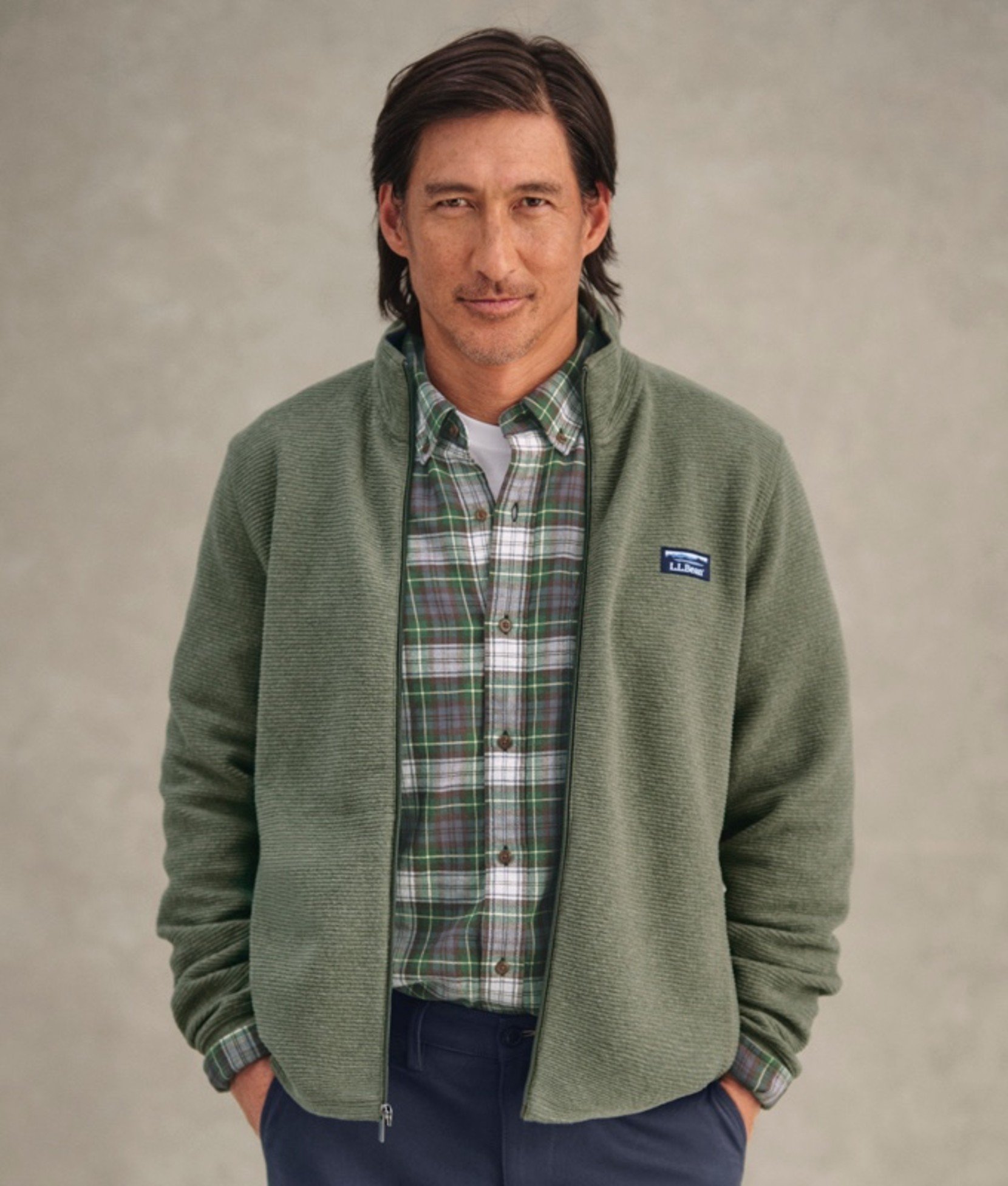 A man wearing an unzipped green fleece jacket over green plaid shirt with hands in pockets. 