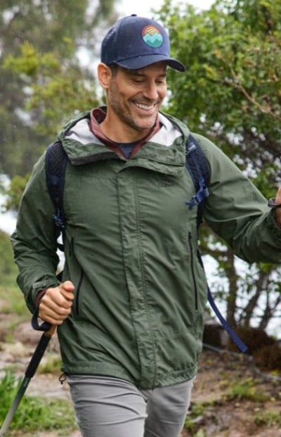 Hiker in a green jacket and cap walks with trekking poles and a backpack on a rainy forest trail.