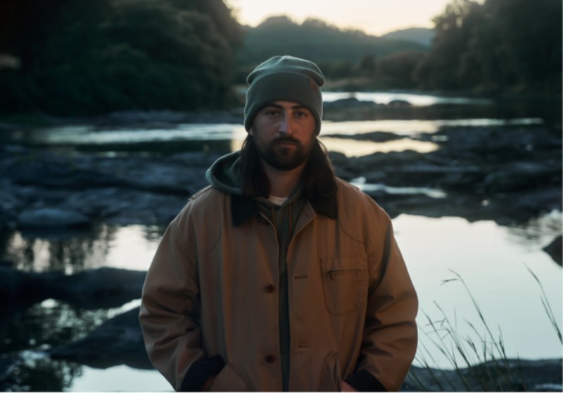 Noah Kahan wearing an L. L. Bean Original Field Coat and beanie stands by a rocky river at dusk.