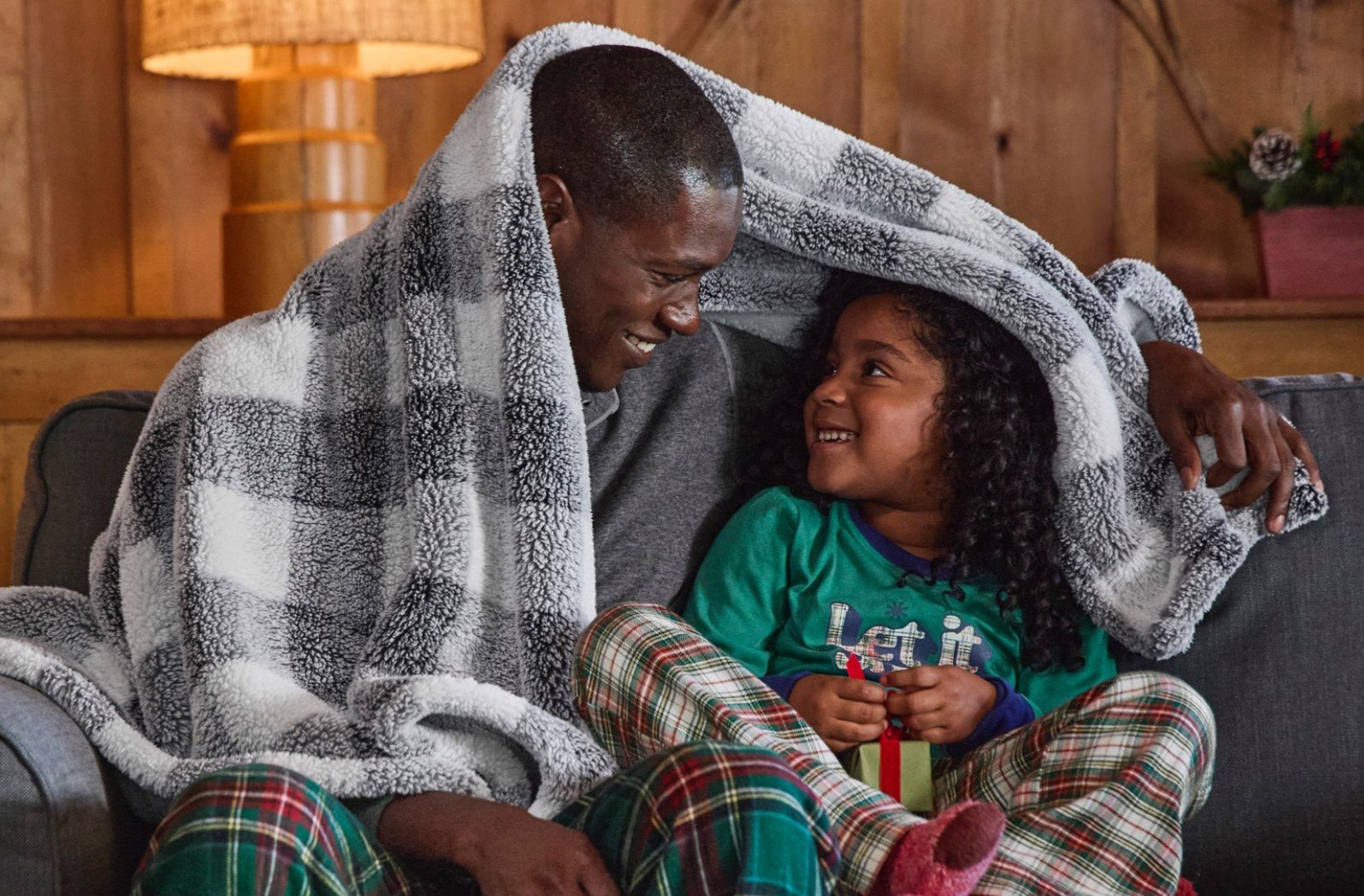 Two people in pajamas sit on a couch, sharing a checkered, cozy, plush fleece throw, with a lamp and wooden wall behind them.