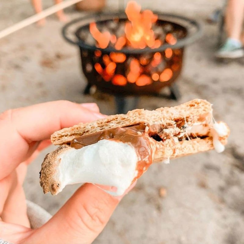 A close-up of a a s'more with a firepit in the background.