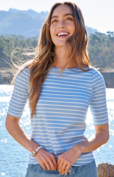 A woman with long hair in a blue and white striped shirt stands near a body of water with trees and mountains in the background.