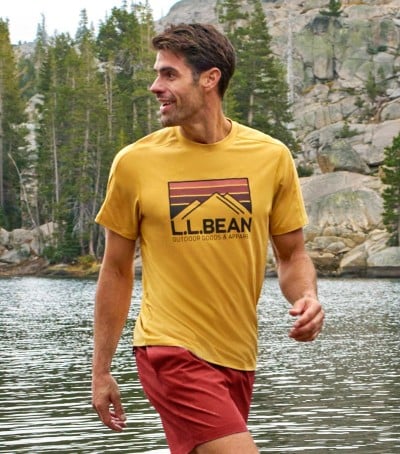 Person in yellow L.L.Bean T-shirt walks along lakeside with trees and rock formations behind.