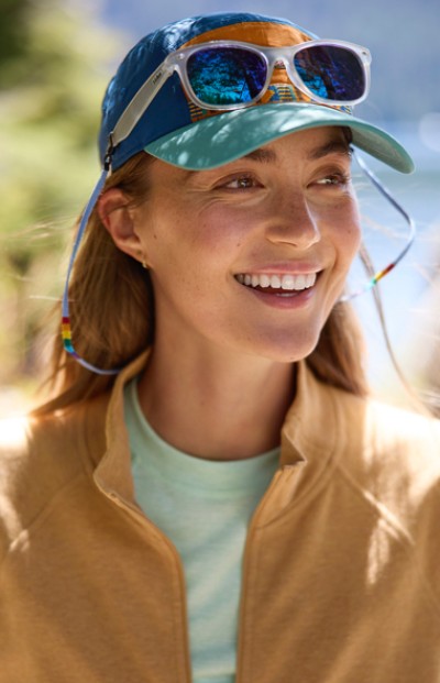 A woman, wearing sunglasses on their head and a cap, stands against a blurred natural backdrop.