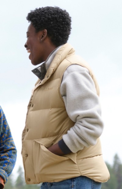 A woman is wearing a beige vest over a grey sweater. The background is not visible, focusing solely on the individual’s attire.