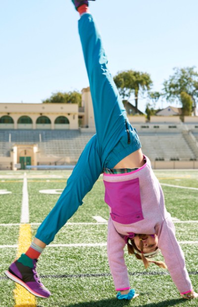 A young girl in a pink top and blue pants performs a cartwheel on a sunny football field.