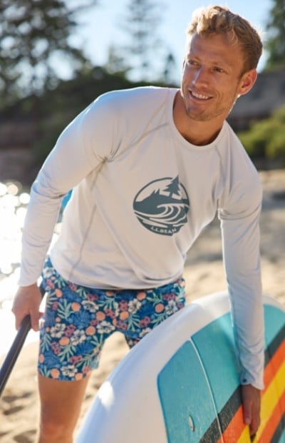 A man in a white long sleeve shirt and floral shorts holds a paddleboard on a sunny beach.
