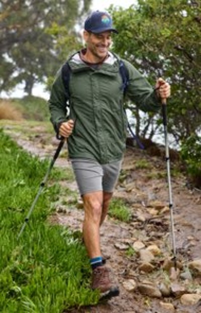 Hiker wearing a green jacket and shorts walks along a muddy trail using trekking poles surrounded by greenery.