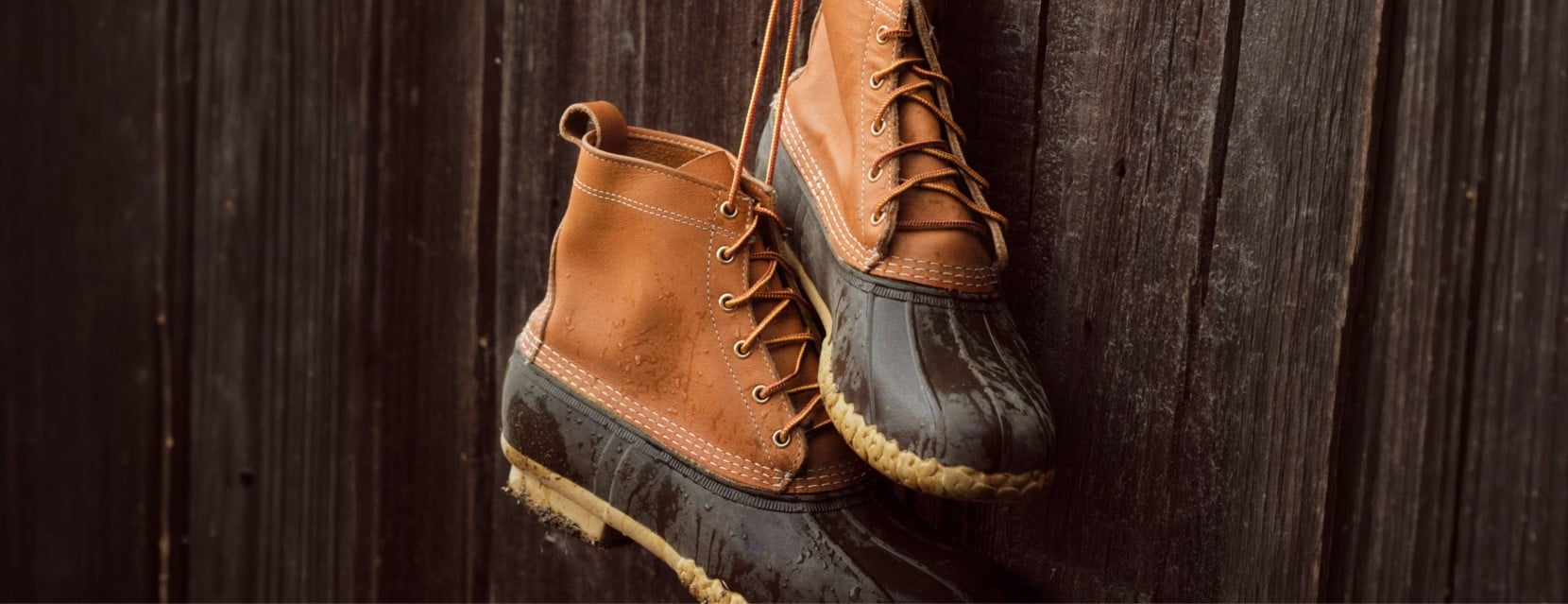 Pair of original L. L. Bean boots hang by their laces against a dark wooden wall, dotted with rain droplets.