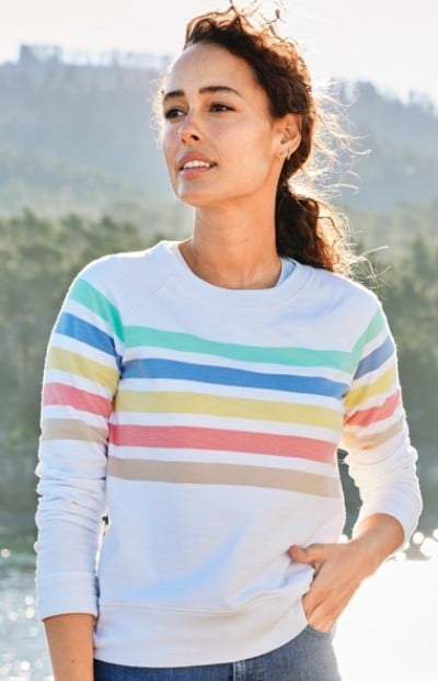 A woman wearing a white sweater with colorful horizontal stripes stands outdoors with a background of trees.