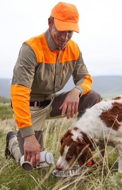 Person kneels on grass in blaze orange and brown outfit, pouring water into a bowl for a brown and white dog.