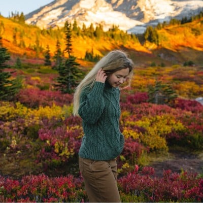 Woman in green sweater and brown pants walks through a colorful meadow with mountains in the background.