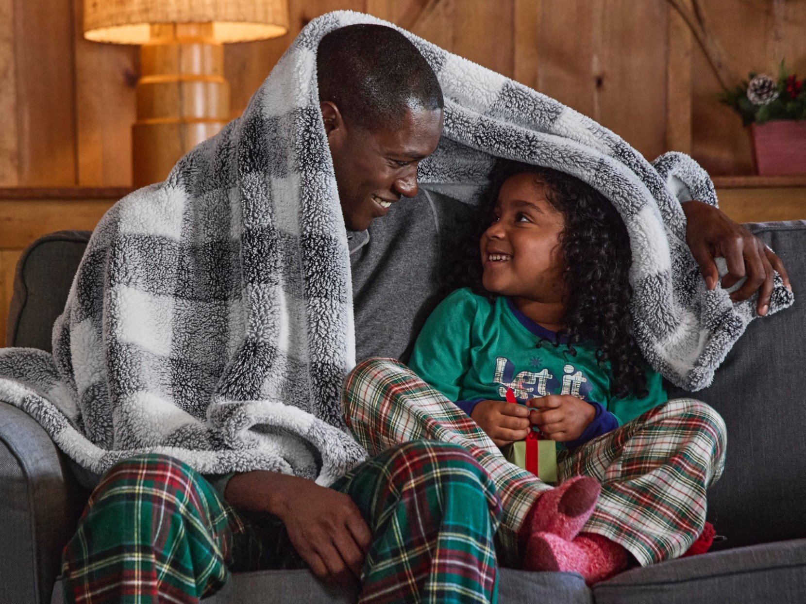 Two people in pajamas sit on a couch, sharing a checkered, cozy, plush fleece throw, with a lamp and wooden wall behind them.