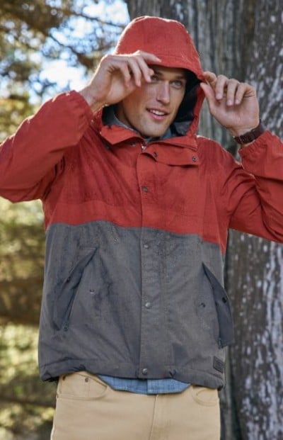 Person adjusts the hood of a redâ€‘andâ€‘gray rain jacket while standing outdoors near a tree.
