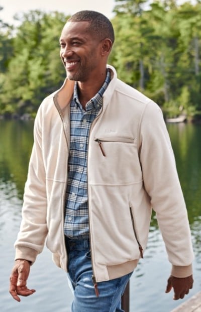 A man wearing a beige jacket and blue plaid shirt stands near a body of water with trees in the background.