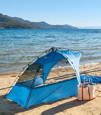 Blue sun shelter and tote bag on sandy lakeshore with calm water and distant mountains. 