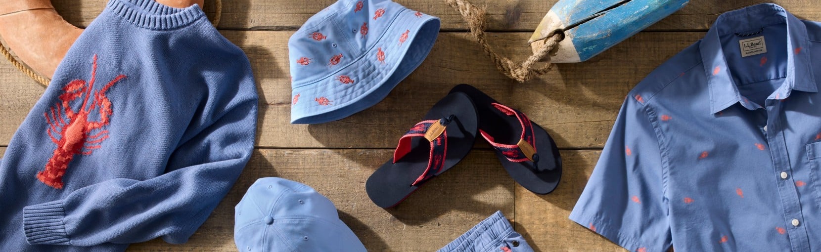 Flat lay of blue lobster-print sweater, shirt, shorts, hat and flip-flops arranged on a wooden surface.