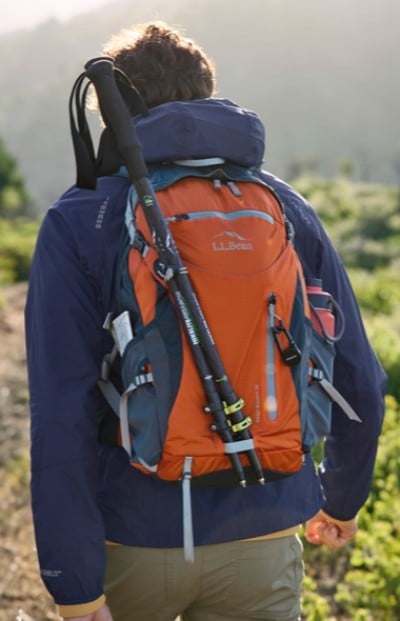 A person wearing a blue jacket and carrying an orange L L Bean backpack with trekking poles attached, walking on a trail in a mountainous area.