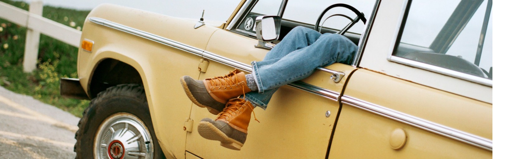 Person resting in a vintage yellow car with legs hanging out the window, wearing jeans and L. L. Bean boots.