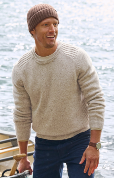 A man wearing a beige sweater, blue jeans, and brown beanie stands near water holding an oar. 