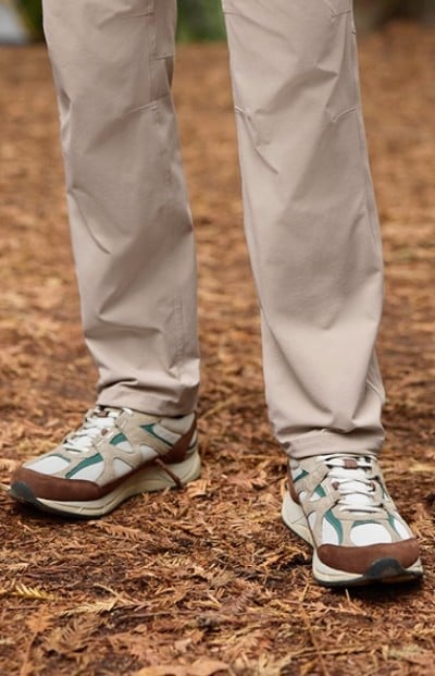 A person wearing beige pants and white, green, and brown hiking shoes stands on a forest floor covered with dried leaves.