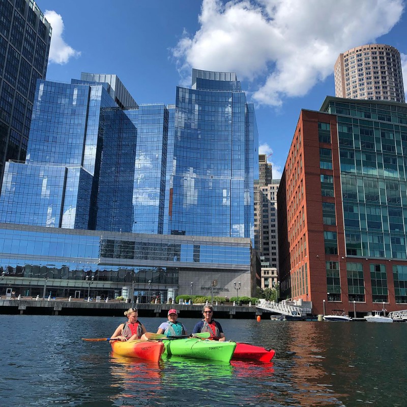 Three kayakers on the water around Fort Point Channel.