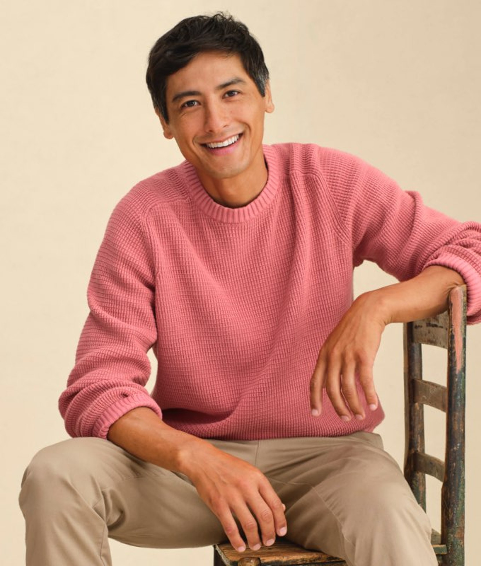 Man seated wearing a pink crewneck sweater and beige pants.