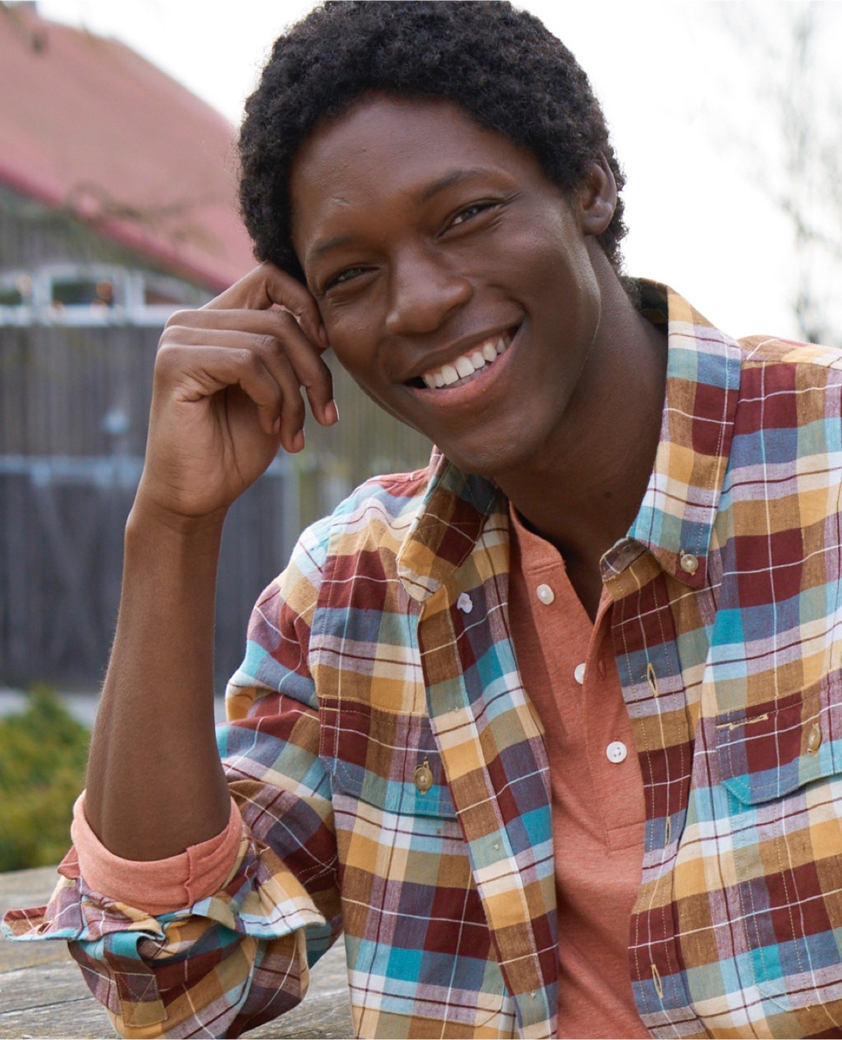 Man in a colorful plaid shirt over orange sits outdoors with hand on head.