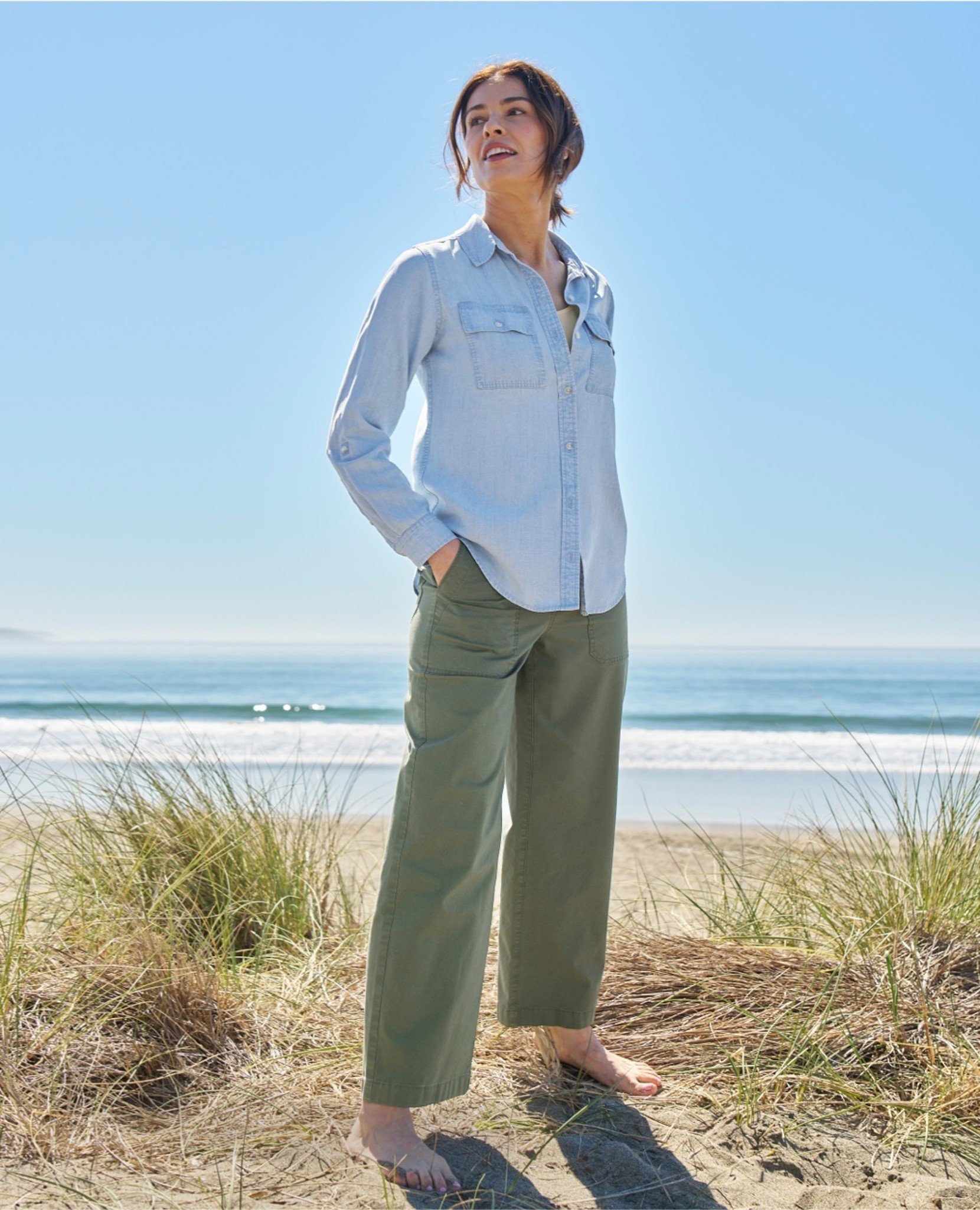 Woman on a sandy beach, wearing a light blue shirt and green pants; ocean in background.
