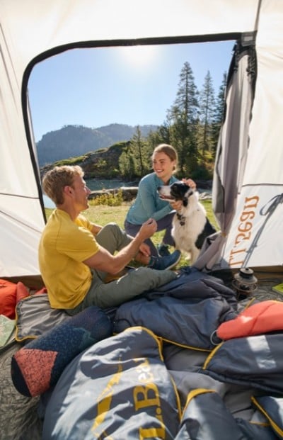 Two people and a dog inside a tent, surrounded by camping gear, with a scenic view of trees, mountains, and a body of water in the background.