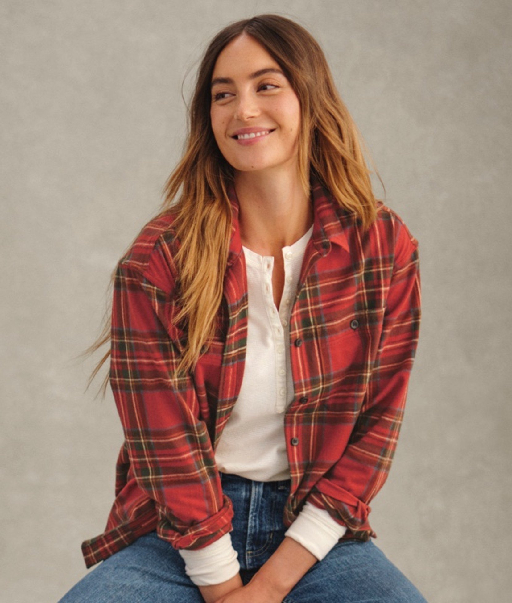 A women in red plaid flannel over white henley and blue jeans, seated.