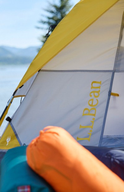 A yellow L L Bean tent is set up with a background of water and hills, suggesting a scenic camping spot. 