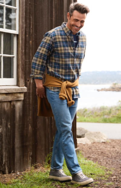 A man in a plaid shirt and jeans stands by a wooden building, face obscured, holding gloves, with a scenic backdrop.