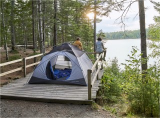 A couple sitting on a fence looking out at the water, their tent set up on a platform behind them.