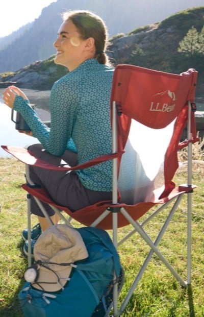A woman in a blue patterned jacket sits on a red L L Bean chair by a lake, holding a cup. A backpack is on the ground beside the chair.