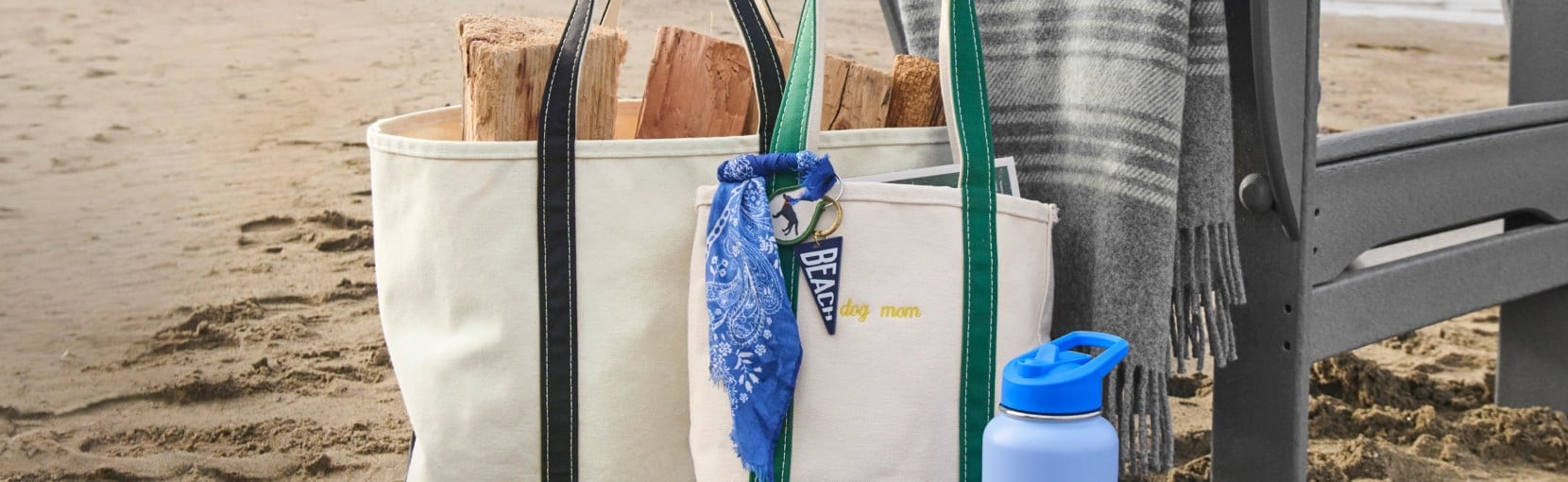 Canvas tote bags filled with firewood sit on a sandy beach beside a chair, a gray blanket, and a blue water bottle.