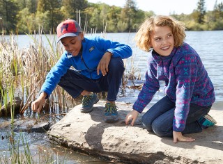 A boy and girl looking into the water from a big rock by a lake.