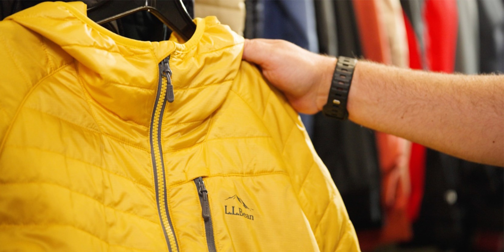 Hand holding yellow L L Bean puffer jacket with hood, surrounded by other jackets.