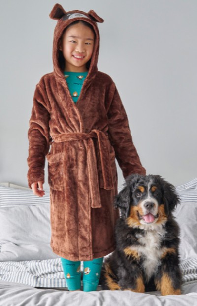 A child in a L L Bean robe stands beside a Bernese Mountain Dog on a bed. 