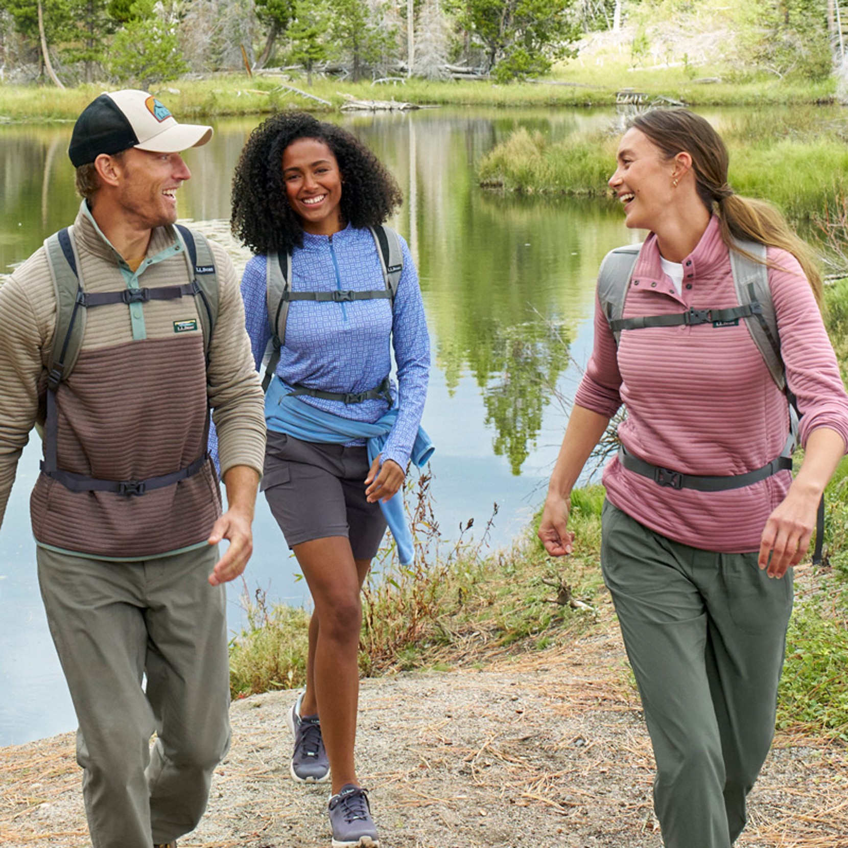 Three people wearing backpacks walk along a lakeside trail, with trees, water, and grassy shore in the background.
